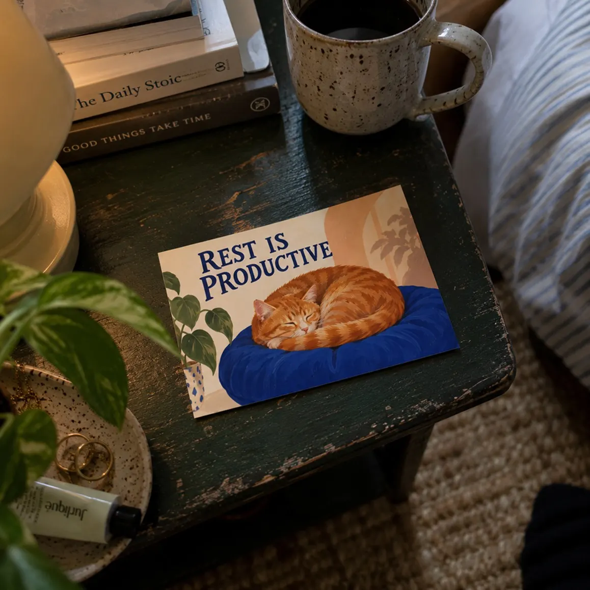 Printed 'Rest is productive' postcard with a sleeping cat on a dark bedside table