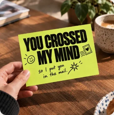 A hand holding a bright yellow-green postcard that reads, ‘You crossed my mind, so I put you in the mail.’